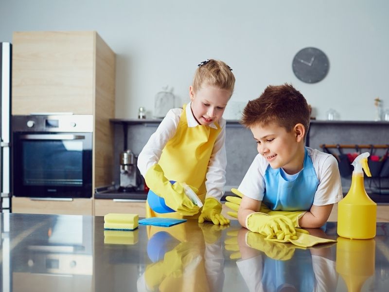 vado kids pflegemittel ein Mädchen und ein junge mit Gummihandschuhen und Putzmitteln wienern eine Edelstahlküchenplatte